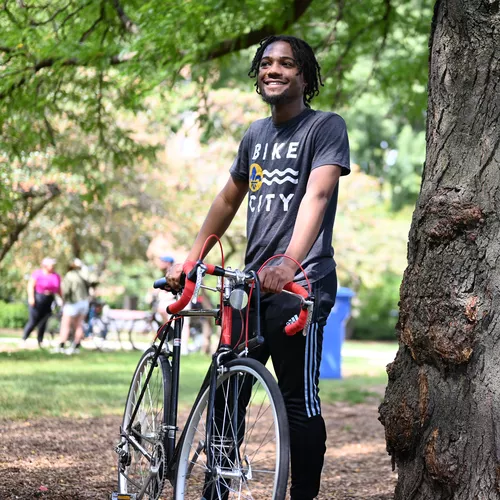 A man stands smiling in park under tree cover, holding his bike. He has dark skin with
            straight neck length dreads and is wearing black track pants and a grey shirt that says
            'Bike City' over some of the symbols of St Louis. The bike is black with red drop
            handlebars.