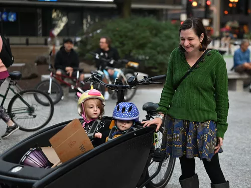 A woman stands smiling looking down at her two young children in a bucket cargo bike
            standing in a downtown plaza. She is wearing a green sweater and a patterned blue and
            yellow skirt, and has neck length brown hair and light skin. Two children sit in the
            black Urban Arrow cargo bike, with a young girl on the left wearing a unicorn helmet and
            a very young boy on the right wearing a blue helmet with white polka dots.
            A cardboard sign and a drum are visible in the bike bucket.