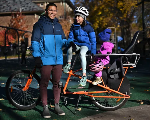 A man stands in front of an orange long-tail bike, with two of his kids sitting on the
            bike, on an autumn day with orange and yellow trees and a playground in the background.
            The man is wearing a blue jacket and red pants with black gloves, and has light brown
            skin, short black hair, and clear glasses. His older daughter is sitting on some bars on
            the bike and smiling at her Dad, while wearing a coat and a helmet over a grey beanie.
            The younger daughter is sitting in a purple sweater onesie and a blue beanie and is
            looking away from camera.