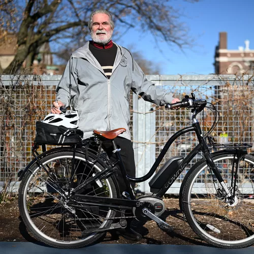 An older man stands smiling with his black ebike in front of a metal fence of Chicago's
            606 trail. He is wearing a silver jacket (with the Bike Lane Uprising logo on it) over a
            dark grey sweater with white stripes, black jeans, and black shoes, and has light skin
            and grey hair and a medium length grey beard. His bike is a black step-through Electra
            townie ebike, with a brown leather seat and swept back handlebars, with a battery and
            Bosch motor visible.